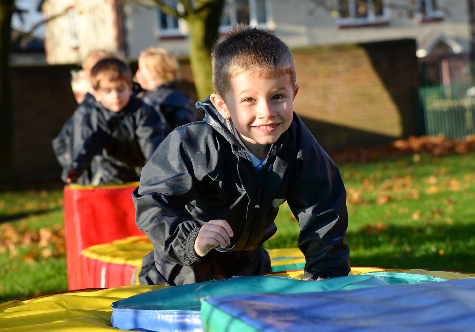 A smiling boy in a dark jacket plays on colorful soft play equipment outdoors.  Other children are blurred in the background.  It's a sunny day.
