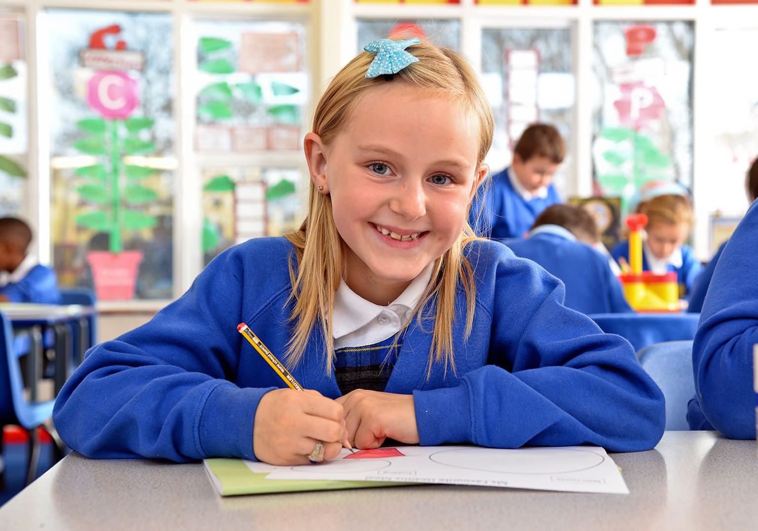 A smiling girl in a blue school uniform sits at a desk, writing with a pencil.  Other children are blurred in the background of a classroom.
