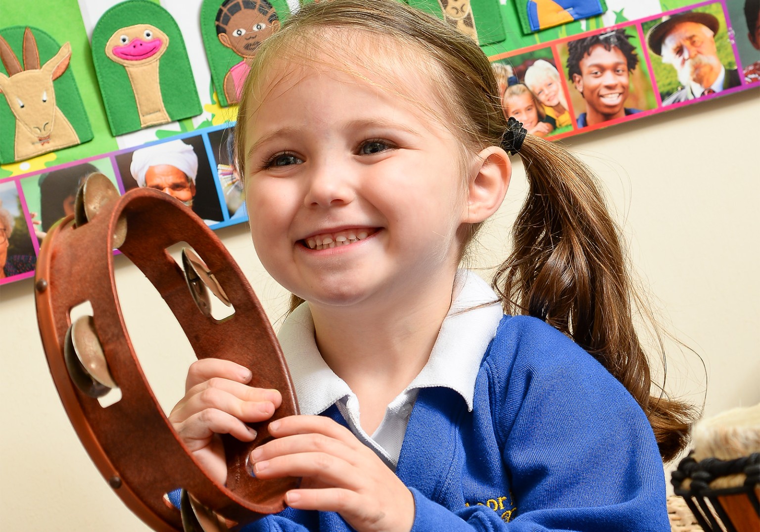 A smiling girl with pigtails holds a tambourine.  A classroom wall with diverse faces and felt animals is in the background.
