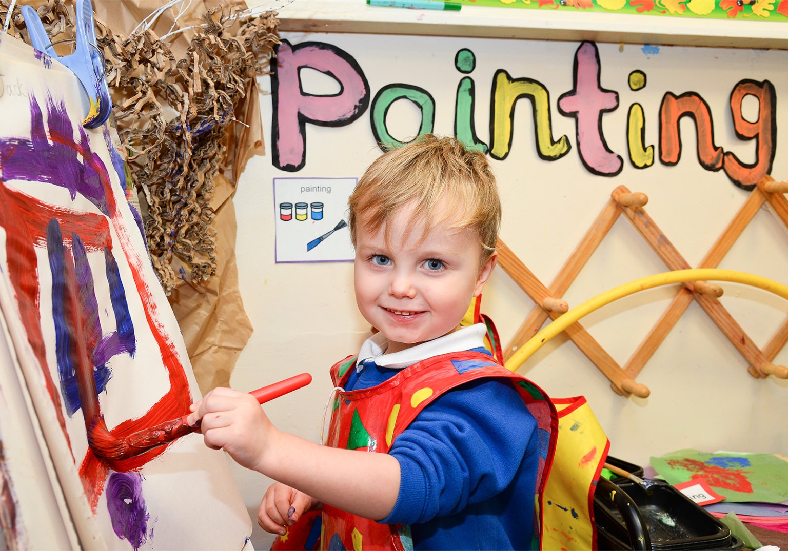 A young boy is painting a picture on an easel. He's wearing a colorful painting smock and smiling at the camera. The word "Painting" is displayed on the wall above him, along with some art supplies.