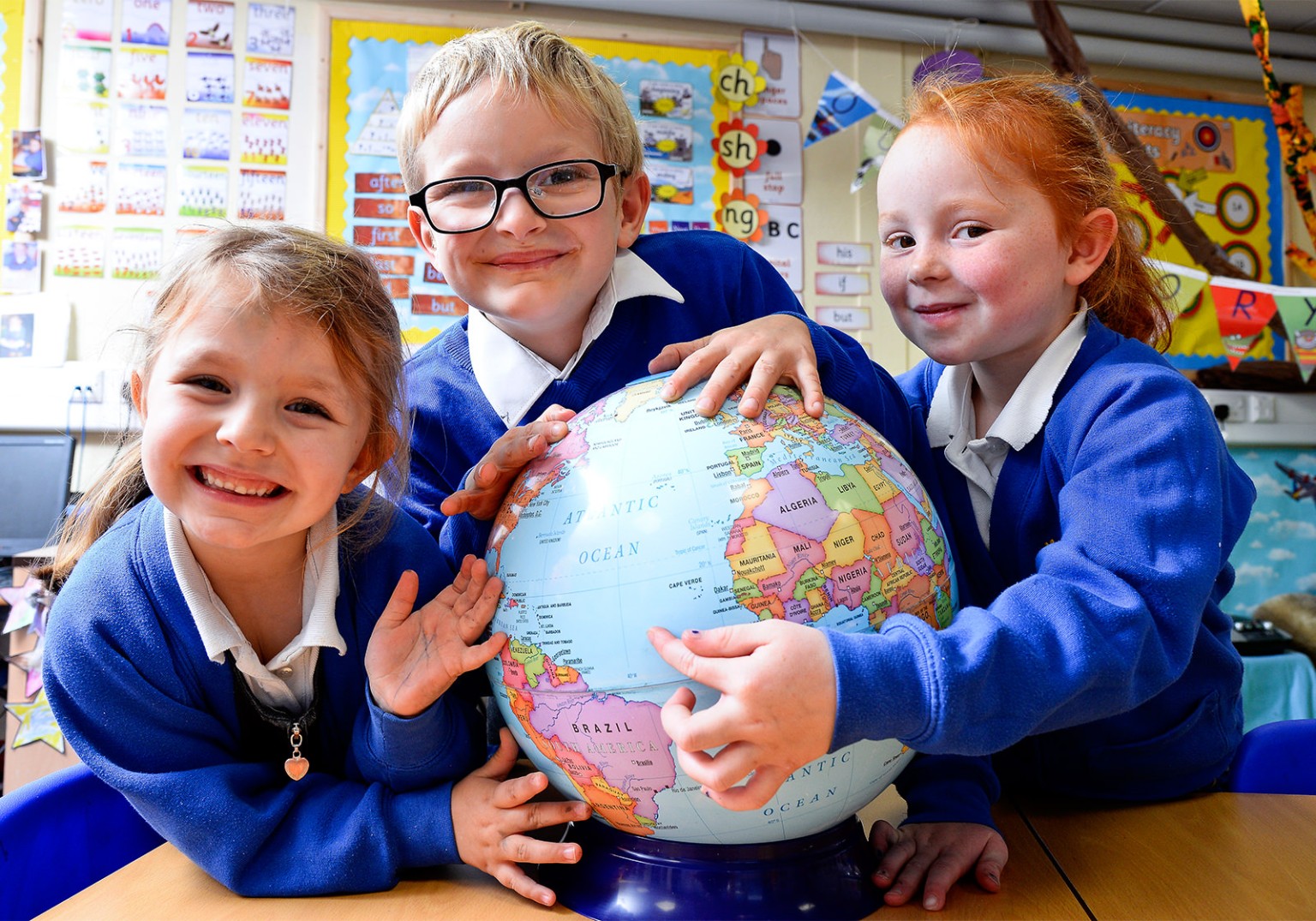 Three young children in blue school uniforms are surrounding a globe, each touching it.  They appear to be in a classroom.
