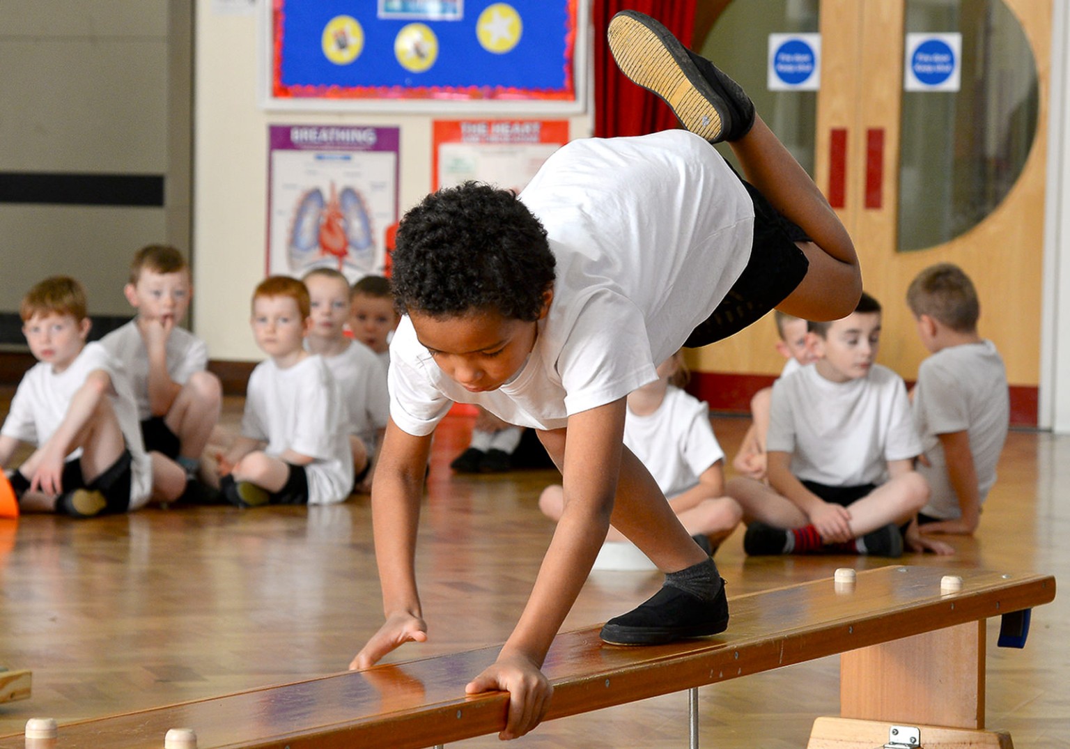 A young boy in a white t-shirt performs a gymnastic move on a low wooden beam. Other children in white shirts sit and watch in a school gymnasium.