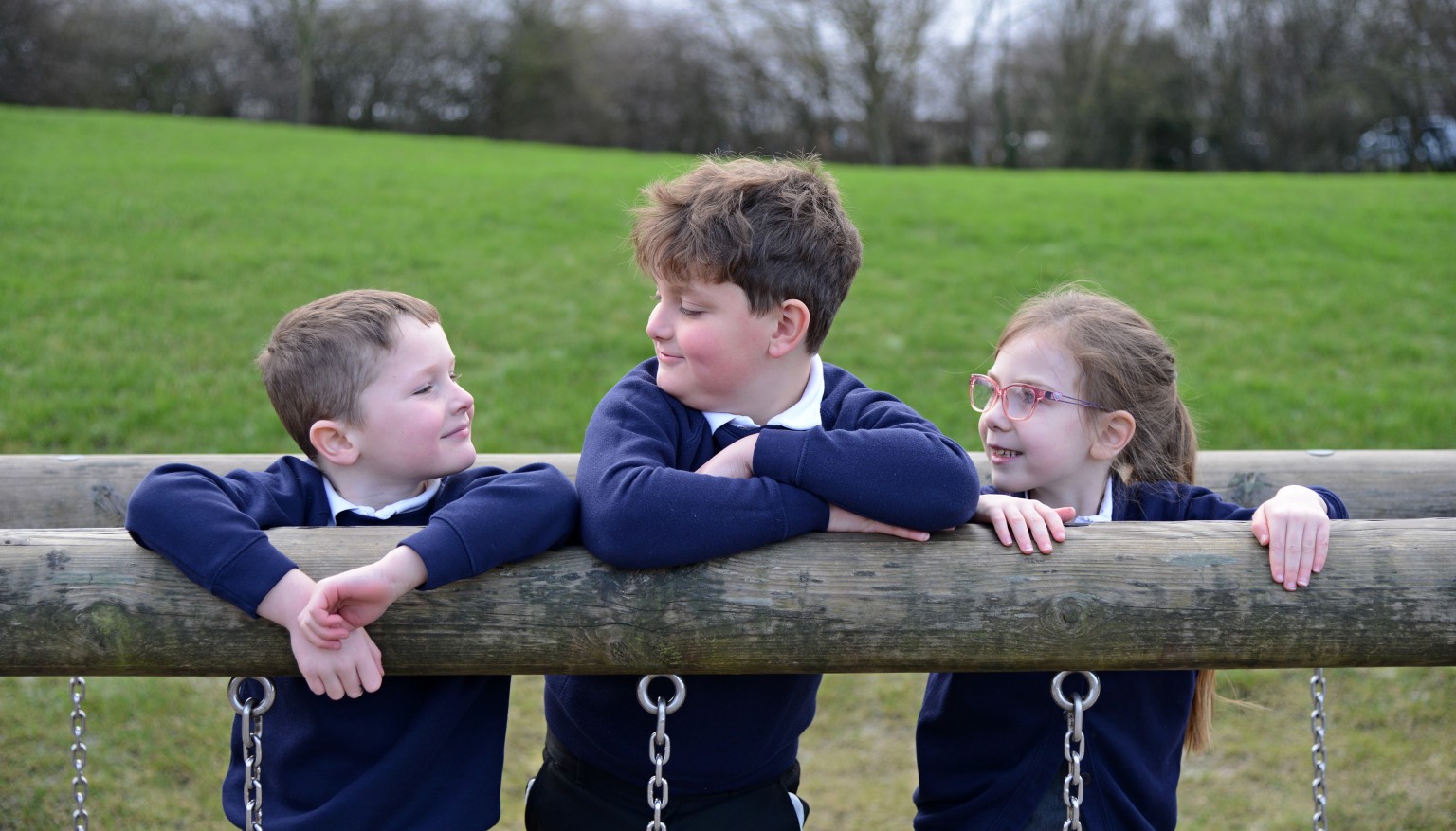 Three children smiling at each other leaning on a trim trail 