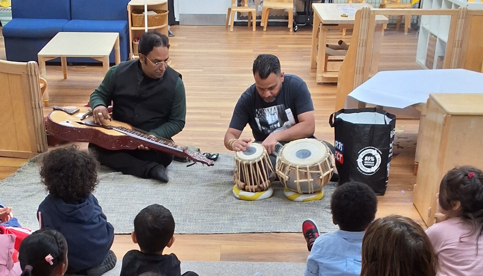 2 Men sitting at the front of a group of children playing the dohl and a stringed instrument.