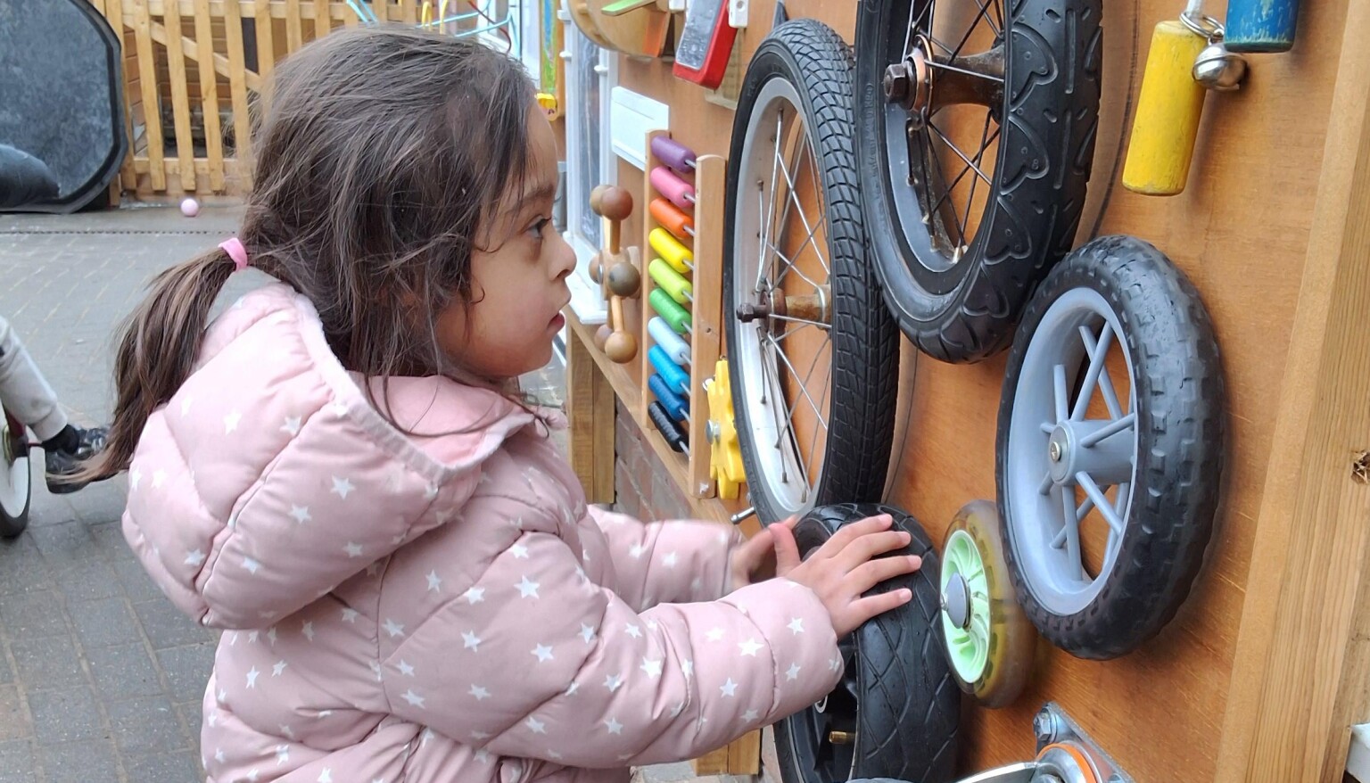 A young child outside waering a coat. They are using their hands to move interlocking wheels attached to a wooden board.