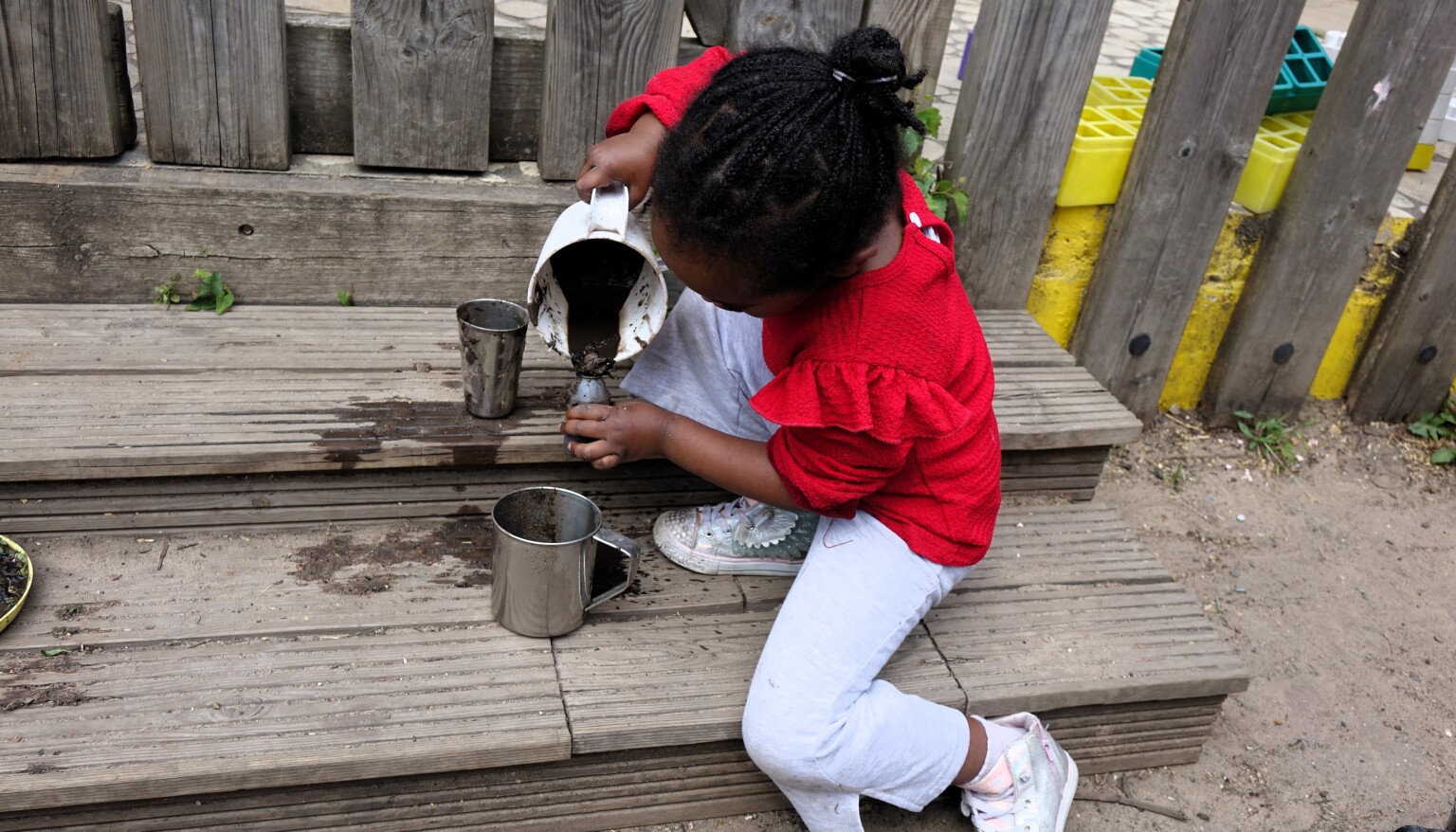 A young child sitting on garden steps and using a jug to pour a watery mu mixture into a metal cup.