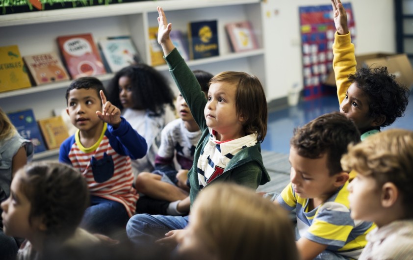 A classroom of preschool-aged children sits on the floor.  Several children have their hands raised, likely participating in a lesson or asking questions. Bookshelves filled with children's books line the wall in the background.
