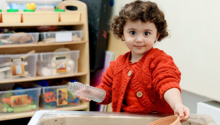 A toddler with curly brown hair, wearing a red cardigan, plays in a sensory bin at a daycare.  She holds a plastic bottle and a small bowl, interacting with the water in the bin.  A storage shelf is visible in the background.
